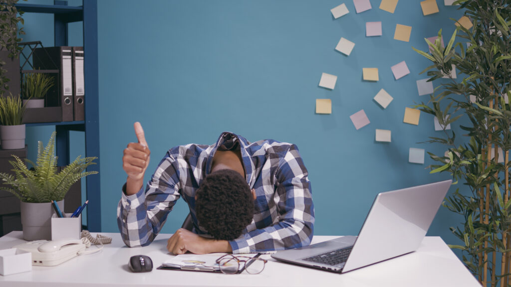 Travailleur épuisé posant la tête sur son bureau et faisant un signe de pouce levé et se sentant fatigué.